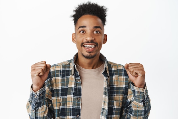 Kostenloses Foto hoffnungsvoller afroamerikanischer mann, der mit aufregung schaut, die fäuste begeistert ballt, auf den fernsehbildschirm schaut, das spiel sieht, nach dem team sucht, ergebnisse vorwegnimmt, auf weiß wetten.