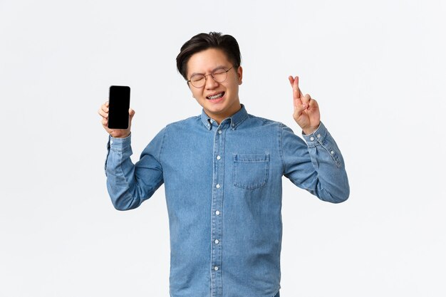 Hoffnungsvoller asiatischer Typ mit geschweiften Augen und gekreuzten Fingern viel Glück beim Zeigen des Smartphone-Bildschirms
