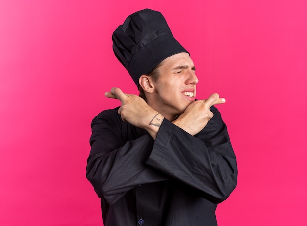Hoffnungsvoller junger blonder männlicher Koch in Kochuniform und Mütze, der die Hände gekreuzt hält und mit geschlossenen Augen viel Glück macht