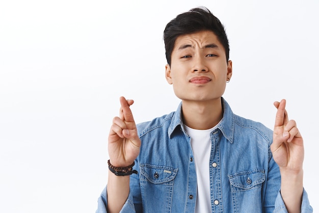 Hoffnungsvoller und intensiver junger asiatischer mann bemüht sich um wunschdenken, kreuzen die finger für viel glück, flehende träume werden wahr, beten alles in ordnung, weiße wand.