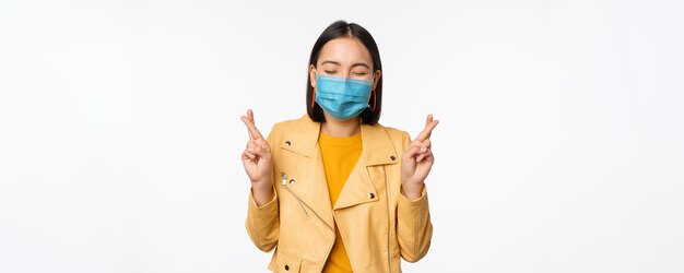 Hoffnungsvolles asiatisches Mädchen in medizinischer Gesichtsmaske kreuzt die Finger und wünscht sich, dass sie für etwas betet, das mit Vorfreude auf weißem Studiohintergrund steht