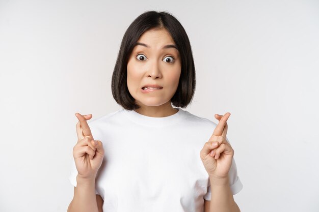 Hoffnungsvolles asiatisches Mädchen kreuzt die Finger und wünscht sich etwas, das im T-Shirt auf weißem Hintergrund steht