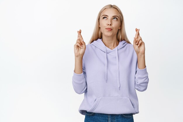 Hoffnungsvolles blondes Mädchen kreuzt die Finger für viel Glück und schaut in die obere linke Ecke, betet, macht den Traum wahr, steht auf Weiß