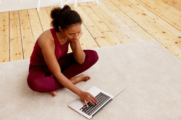 Hohe Winkelansicht der jungen afroamerikanischen Frau, die Leggings und oben sitzt, die mit gekreuzten Beinen auf der Matte vor offenem Laptop sitzen, WiFi verwenden, nach Tutorial suchen, Yoga zu Hause praktizieren