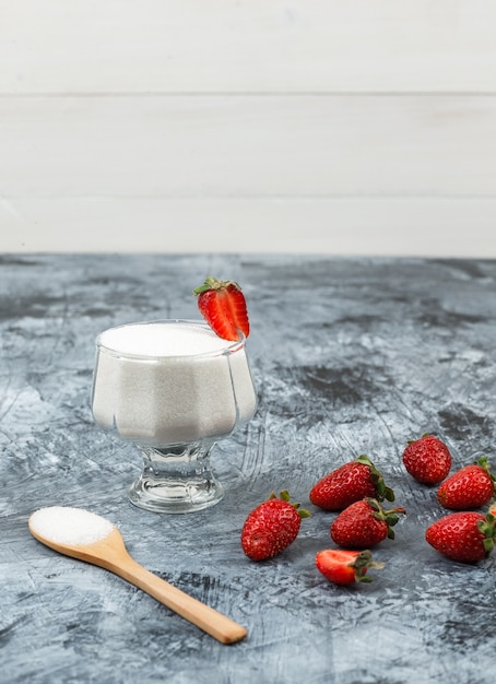 Hohe Winkelansicht einer Glasschale Joghurt auf Weiden-Tischset mit Holzlöffel und Erdbeeren auf dunkelblauem Marmor und weißem Holzbretthintergrund. vertikaler freier Speicherplatz für Ihren Text