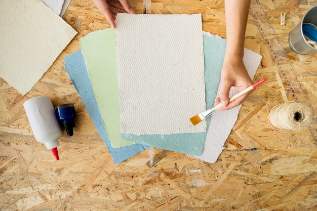 Hohe Winkelsicht der Hand einer Frau unter Verwendung des Malerpinsels über handgemachten Papieren