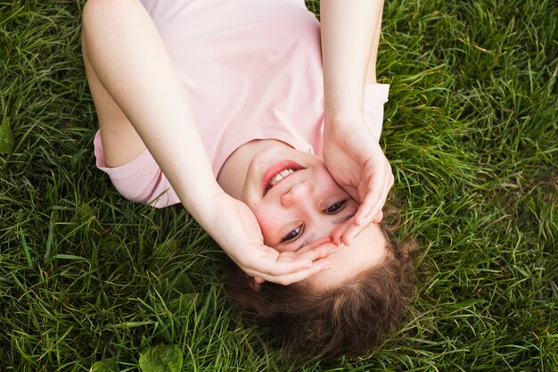 Hohe Winkelsicht des lächelnden Mädchens liegend auf Gras und ihre Augen abschirmend
