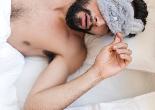 Hohe Winkelsicht eines hemdlosen Mannes, der auf Bett mit einer Augenmaske schläft