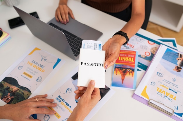 Kostenloses Foto hoher blickwinkel auf den schreibtisch eines reisebüros mit menschen, die am laptop arbeiten und einen reisepass in der hand halten