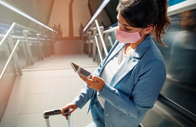 Hoher Winkel der Frau mit medizinischer Maske und Gepäck unter Verwendung des Smartphones am Flughafen während der Pandemie