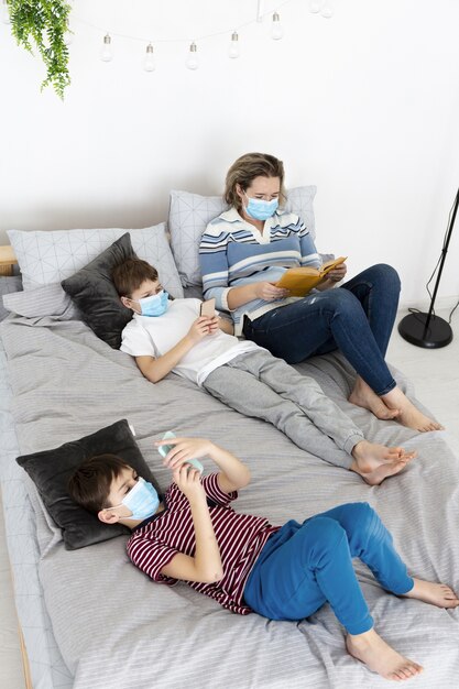 Hoher Winkel der Kinder im Bett mit medizinischen Masken und Mutter, die ein Buch lesen