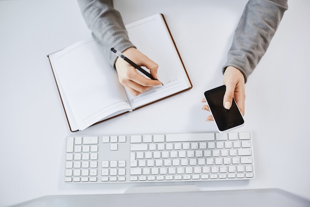 Hoher Winkelschuss der beschäftigten fleißigen Frau, die Smartphone hält. Kurzer Schuss von Frauenhänden, die Notizen im Planer machen und in der Nähe von Tastatur und Computer sitzen, bei der Arbeit sind und mit Teamkollegen arbeiten