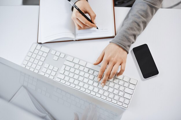 Hoher Winkelschuss der Frauenhände, die auf Tastatur tippen. Konzentrierte Studentin, die an ihren Hausaufgaben am Computer arbeitet, Notizen macht, auf einen wichtigen Anruf vom Chef wartet und einen Blick auf das Smartphone wirft.