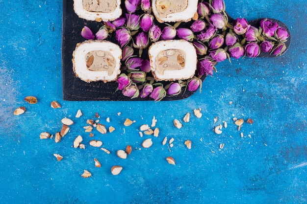 Holzbrett mit süßen Köstlichkeiten und getrockneten Rosen auf blauem Tisch.