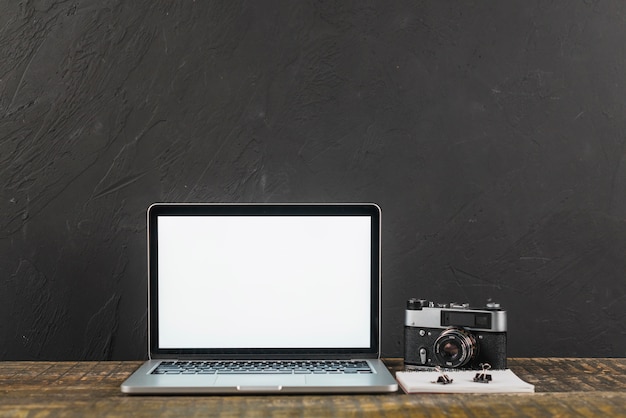 Kostenloses Foto holztisch mit laptop des leeren bildschirms und retro- fotokamera auf schwarzem hintergrund
