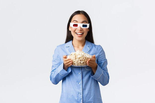 Home Freizeit, Übernachtung und Pyjama-Party-Konzept. Begeistertes asiatisches Mädchen im Pyjama und 3D-Brille, das eine Schüssel Popcorn hält und amüsiert lächelt, als es Premiere im Fernsehen sieht und den Filmabend genießt