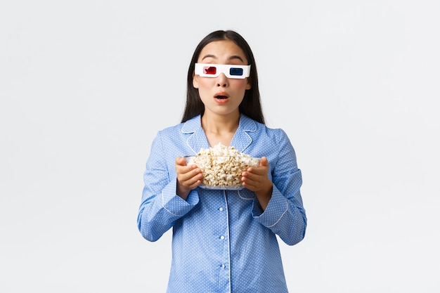 Home Freizeit, Übernachtung und Pyjama-Party-Konzept. Erstauntes und beeindrucktes asiatisches Mädchen im Schlafanzug, das Thriller ansieht, eine Schüssel Popcorn hält und eine 3D-Brille trägt, in Ehrfurcht in den Fernseher starrt.