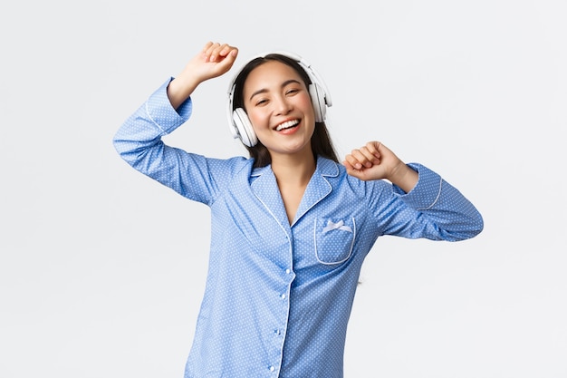 Home Freizeit, Wochenenden und Lifestyle-Konzept. Fröhliches, glückliches asiatisches Mädchen im Pyjama, das Spaß hat, zu Musik in Kopfhörern tanzt, am freien Tag Lieblingslied hört, fröhlicher weißer Hintergrund steht