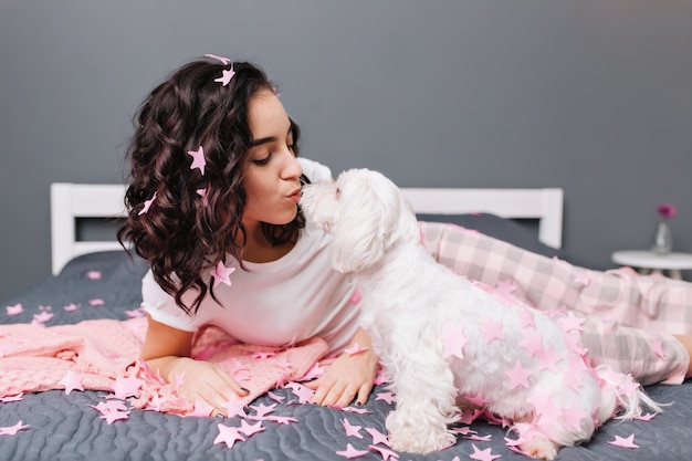 Home glückliche Momente mit Haustieren der jungen schönen Frau im Pyjama mit geschnittenem brünettem lockigem Haar in rosa Lametta auf Bett in moderner Wohnung. Schönes hübsches Modell, das zu Hause mit weißem kleinen Hund chillt