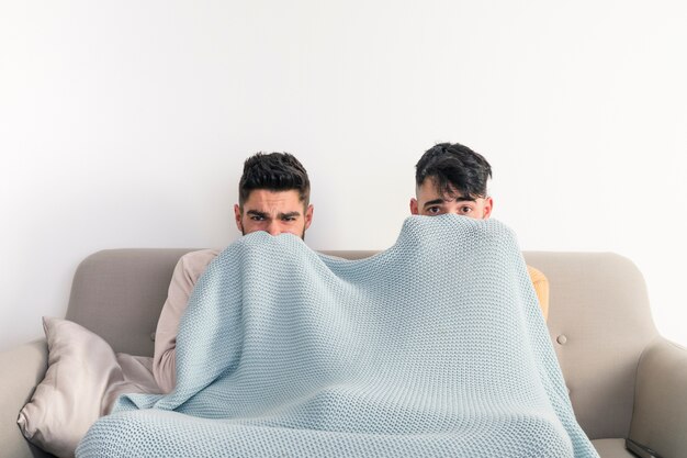 Homosexuelle Paare, die auf dem Sofa bedeckt ihren Mund mit blauer Decke beim Fernsehen sitzen