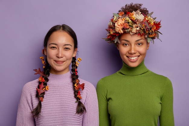 Horizontale Ansicht von fröhlichen Frauen stehen nebeneinander, drücken positive Emotionen aus, verzieren Haare mit Herbstattributen, isoliert über lila Hintergrund