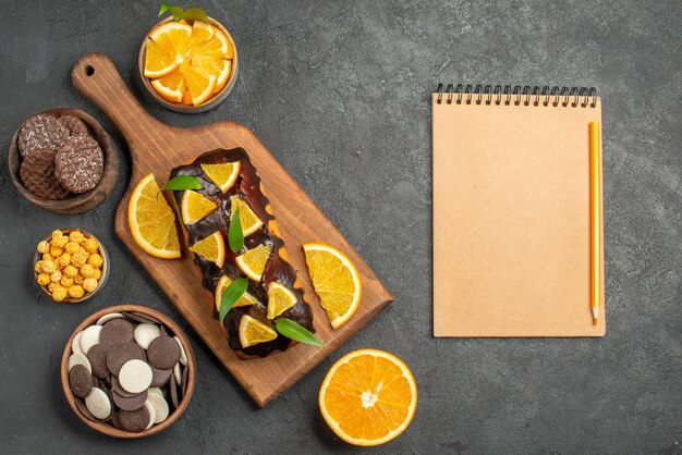 Horizontale Ansicht von leckeren Kuchen schneiden Orangen mit Keksen und Notizbuch auf Schneidebrett auf dunklem Tisch