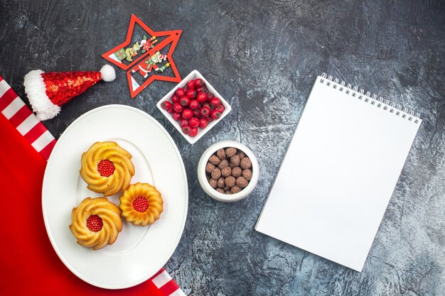 Horizontale Ansicht von Notebook und leckeren Keksen auf einem weißen Teller auf rotem Handtuch und Weihnachtsmann-Hut Cornel und Chocoltes in weißen Töpfen auf dunkler Oberfläche