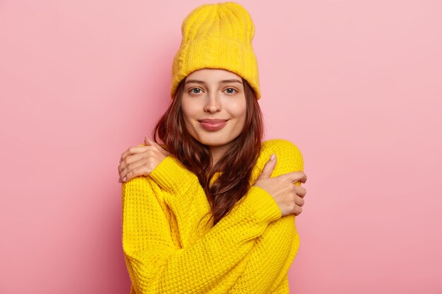 Horizontale Aufnahme der attraktiven jungen Frau umarmt sich, hat dunkles langes Haar, zarten Blick, trägt gelbe Wintermütze und Pullover, posiert gegen rosa Studiohintergrund.