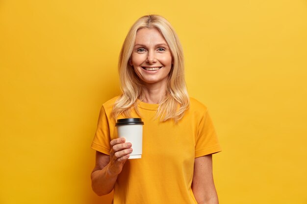 Horizontale Aufnahme der blonden europäischen Frau mit angenehmem Lächeln minimales Make-up hält Wegwerfbecher Kaffee gekleidet in lässigem T-Shirt