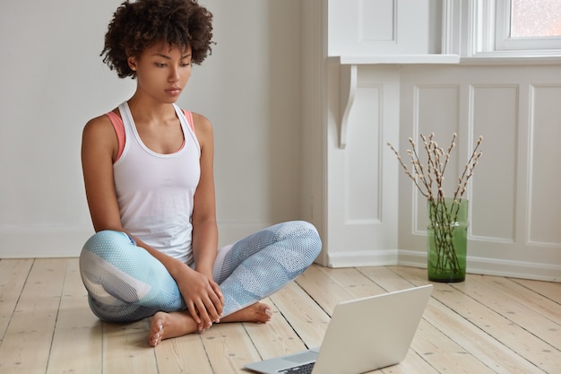 Kostenloses Foto horizontale aufnahme der entspannten dunkelhäutigen frau sitzt in lotus-pose, sieht trainingsvideo, lernt yoga, gekleidet in freizeitkleidung, posiert auf holzboden in leerem raum. technologie- und ruhekonzept