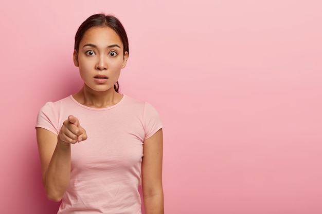 Horizontale Aufnahme der erschrockenen asiatischen Frau zeigt Zeigefinger, fühlt sich verlegen, etwas Seltsames zu bemerken, hat dunkles Haar in Pferdeschwanz gekämmt, trägt rosiges T-Shirt, isoliert auf rosa Wand