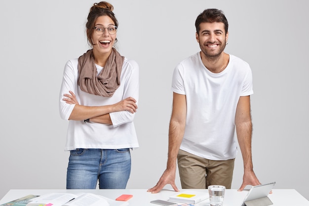 Horizontale Aufnahme der fröhlichen europäischen Frau mit verschränkten Armen, aufgeregtem Blick, modischem Outfit, reagiert auf gutes Ergebnis der Projektarbeit, steht neben Geschäftspartner