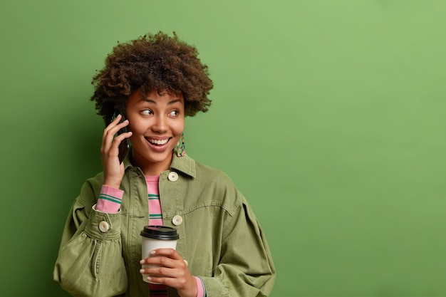 Horizontale aufnahme der hübschen afroamerikanischen frau hat telefongespräch hält smartphone in der nähe von ohrgetränken kaffee zum mitnehmen schaut weg mit sanftem lächeln isoliert über grüner wand kopie raum