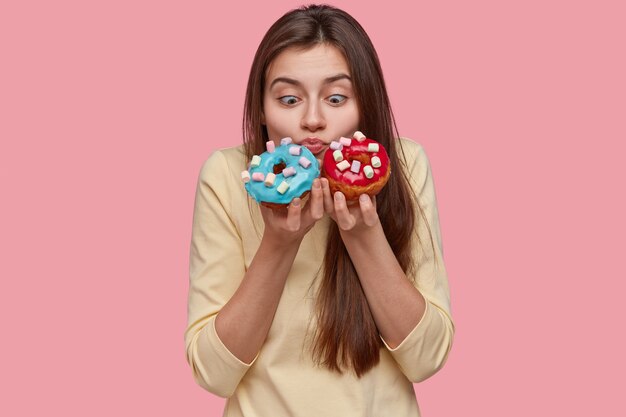 Horizontale Aufnahme der schockierten hübschen europäischen Frau hält blaue und rote Donuts, riecht aromatische Süßwaren
