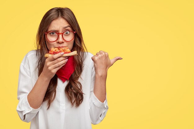 Horizontale Aufnahme der überraschten Frau isst leckeres Stück Pizza, gekleidet in modische Kleidung, zeigt mit dem Daumen an, lädt Sie zur Pizzeria ein, isoliert über gelber Wand. Menschen und Ernährung