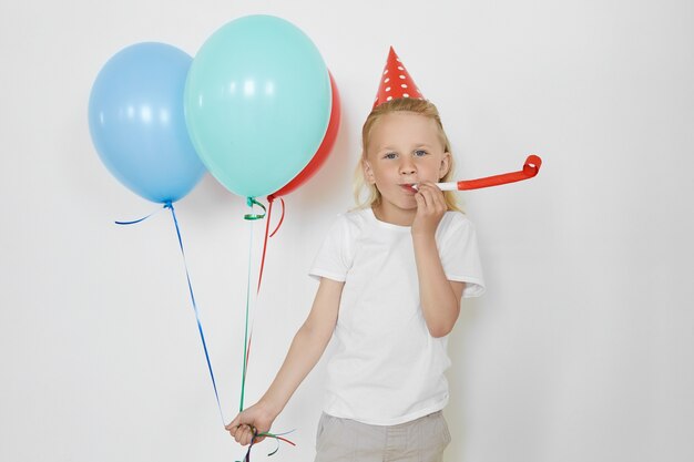 Horizontale Aufnahme des blonden hübschen europäischen Geburtstagskindes, der Freizeitkleidung und roten Kegelhut trägt, Party genießt, Horn bläst, bunte Luftballons hält, mit glücklichem Ausdruck