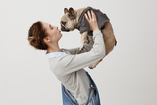 Horizontale Aufnahme des erwachsenen Mädchens in Jeansoveralls, die niedlichen Welpen küssen, während sie ihn in der Luft erhöhen. Junges Mädchen, das in ihren Hund verliebt ist, der im trendigen Pullover gekleidet ist. Anzeige der Zuneigung