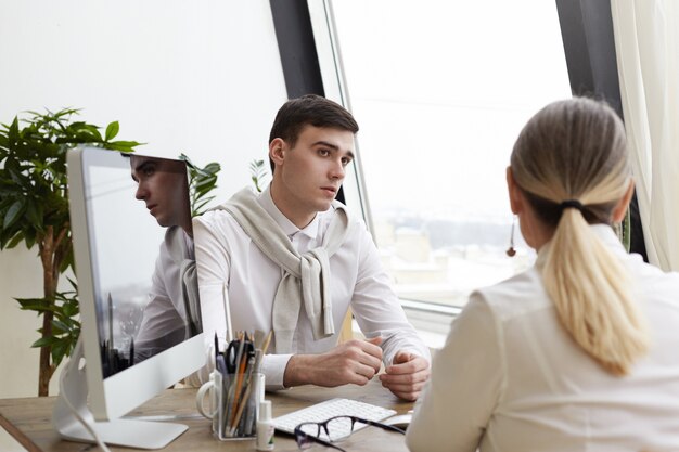 Horizontale Aufnahme des hübschen jungen brünetten Mannrekrutierers, der stilvolle formelle Kleidung trägt, die unerkennbaren reifen weiblichen Berufskandidaten interviewt und ihr im modernen Büroinnenraum aufmerksam zuhört