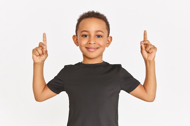 Horizontale Aufnahme des hübschen sportlichen afroamerikanischen Jungen im stilvollen schwarzen T-Shirt, das lokal mit den angehobenen Vorderfingern aufwirft, die Zeigefinger nach oben zeigen und Kopienraum für Ihre Informationen zeigen