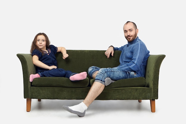 Horizontale Aufnahme des lustigen Schulmädchens, das auf grünem Sofa mit ihrem bärtigen Vater sitzt, der Freizeitkleidung trägt und Kamera mit emotionalen schockierten Ausdrücken gegen weißen Wandhintergrund anstarrt