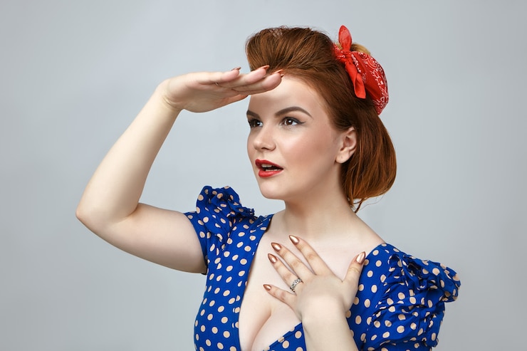 Kostenlose Foto horizontale aufnahme des schönen glamourösen pin-up-mädchens mit hellem make-up und retro-kleid mit tiefem schnitt, das die handfläche an der stirn hält und in die ferne schaut, um etwas zu sehen