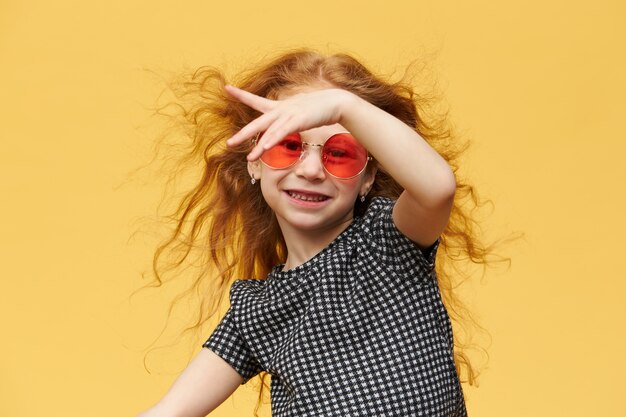 Horizontale Aufnahme des schönen glücklichen trendigen kleinen Mädchens mit dem lockigen Ingwerhaar, das tanzend genießt, mit fröhlichem breitem Lächeln, Sonnenbrille tragend. Musik, Tanz, Spaß und Kinderkonzept