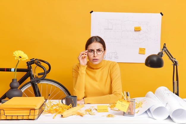 Horizontale Aufnahme einer brünetten Frau sieht aufmerksam aus, hält die Hand an der Brille und versucht, etwas in Fernposen bei unordentlichen Desktop-Arbeiten zum Zeichnen für Architekturprojekte zu sehen.