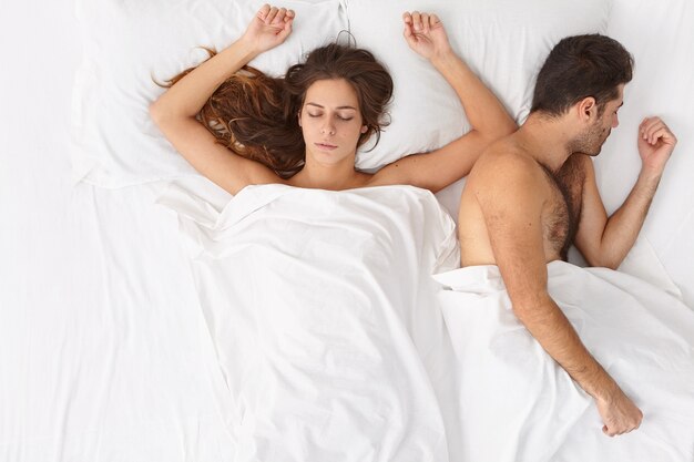 Horizontale Aufnahme einer entspannten verheirateten Frau und eines Mannes, die zusammen im Bett bleiben, einen gemütlichen Morgen und Intimität genießen, einen gesunden Schlaf haben, sich nach leidenschaftlichem Sex ausruhen und unter weißen Laken liegen. Gute Nachtruhe