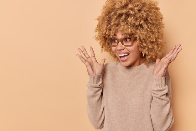 Horizontale Aufnahme einer Frau mit lockigen Haaren exlcaims fühlt sich laut emotional angehoben, die Hände erhoben, trägt einen lässigen Pullover und eine Brille, die über beigem Hintergrund mit Kopierbereich isoliert sind. Menschliche Reaktion