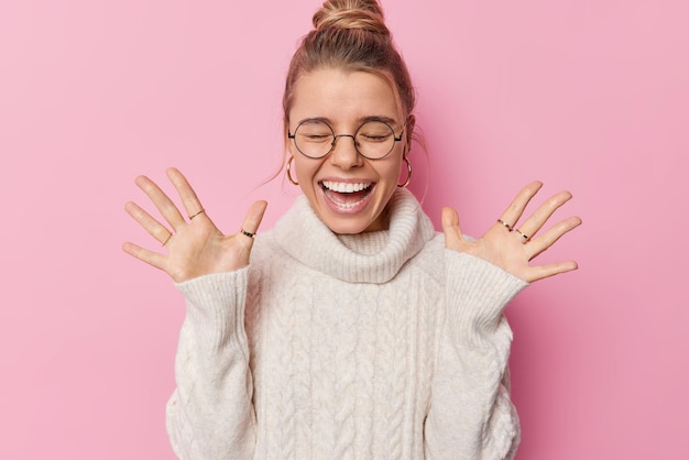 Kostenloses Foto horizontale aufnahme einer glücklichen frau mit gekämmten haaren hebt handflächen, trägt eine runde brille und weißer pullover fühlt sich amüsiert vor rosa hintergrund posiert und hört erstaunliche gute nachrichten, die vor glück schreien