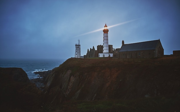 Horizontale Aufnahme einer mysteriösen Stadt auf einer Klippe mit einem weißen eingeschalteten Leuchtturm während der Dämmerung