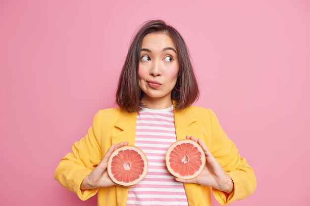 Horizontale Aufnahme einer nachdenklichen asiatischen Frau, die zwei Grapefruithälften hält, die konzentriert sind, trägt stilvolle Kleidung, die über rosafarbener Wand isoliert ist. Natürliche Ernährung und saftiges Zitrusfruchtkonzept.