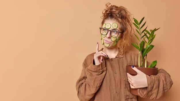 Horizontale Aufnahme einer nachdenklichen jungen Europäerin trägt eine Gurkenmaske zur Feuchtigkeitspflege der Haut auf, trägt eine Brille und Pyjama umarmt Topfpflanze isoliert über braunem Hintergrund mit Leerzeichen