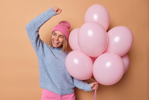 Horizontale Aufnahme einer positiven Europäerin trägt einen Hut, lässiger blauer Pullover, tanzt sorglos und genießt die Geburtstagsfeier, hält einen Haufen aufgeblasener Luftballons, die über braunem Hintergrund isoliert sind. Festlicher Anlass